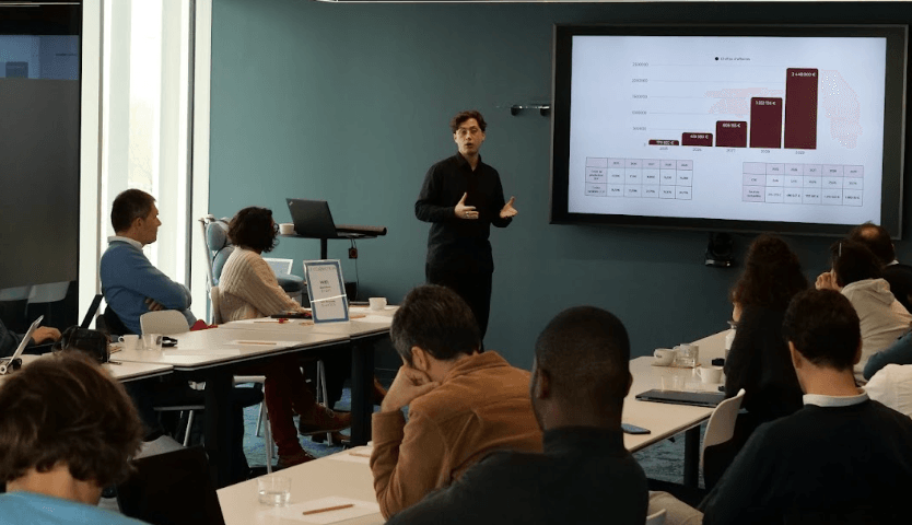 Atelier ou présentation : intervenant devant un écran avec graphiques de chiffre d’affaires, public assis autour de tables.